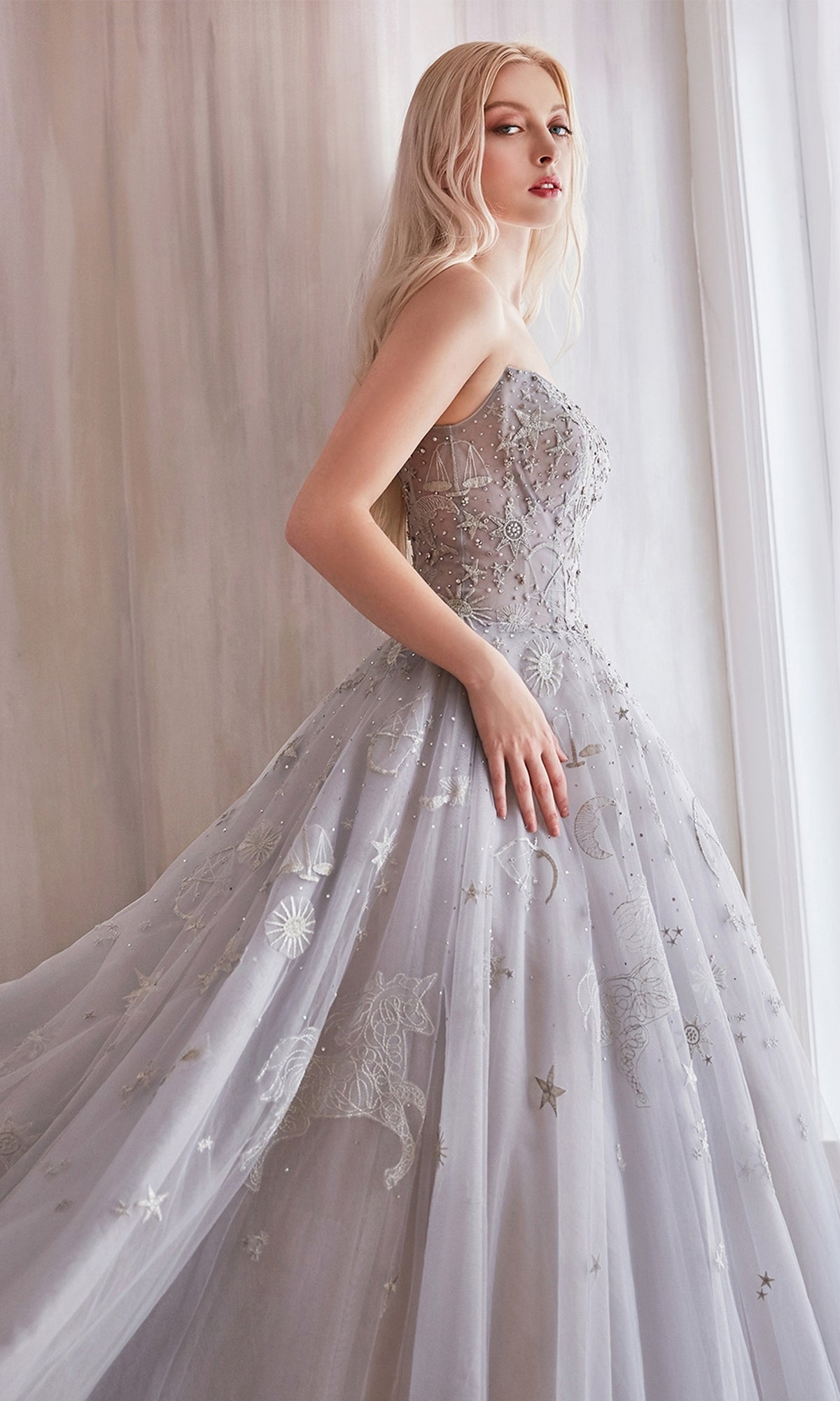 A woman in the Constellation-Print Strapless Prom Ball Gown A0890, featuring star and moon motifs, stands near a softly lit window.
